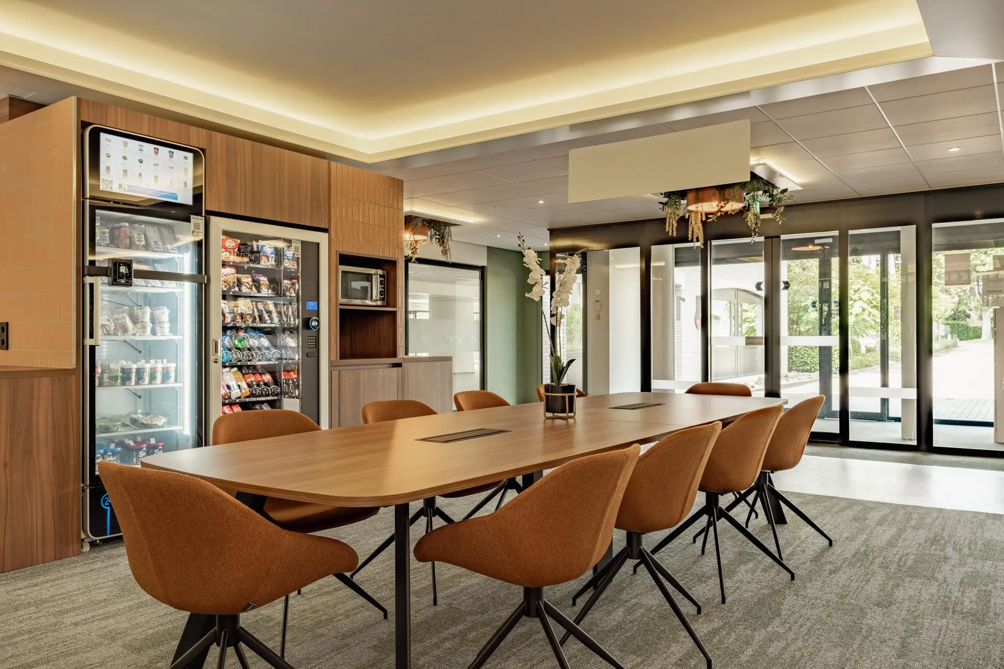 Moderne vergaderruimte met houten tafel, oranje stoelen en een wand met snackautomaten en een magnetron.